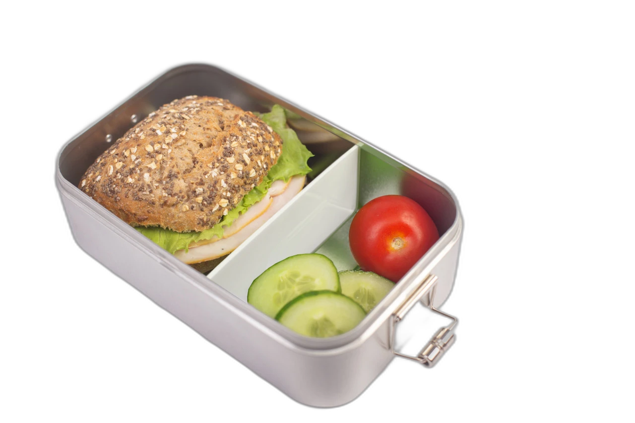 Rechteckige silberfarbene Brotdose mit eingesetzter weißer Trennwand, gefüllt mit einem Körnerbrötchen mit Salat sowie Tomate und Gurkenscheiben, auf weißem Hintergrund fotografiert.