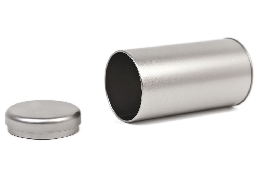 Geöffnete runde Teedose aus silberfarbenem Metall, liegend dargestellt, mit abgenommenem Stülpdeckel daneben.