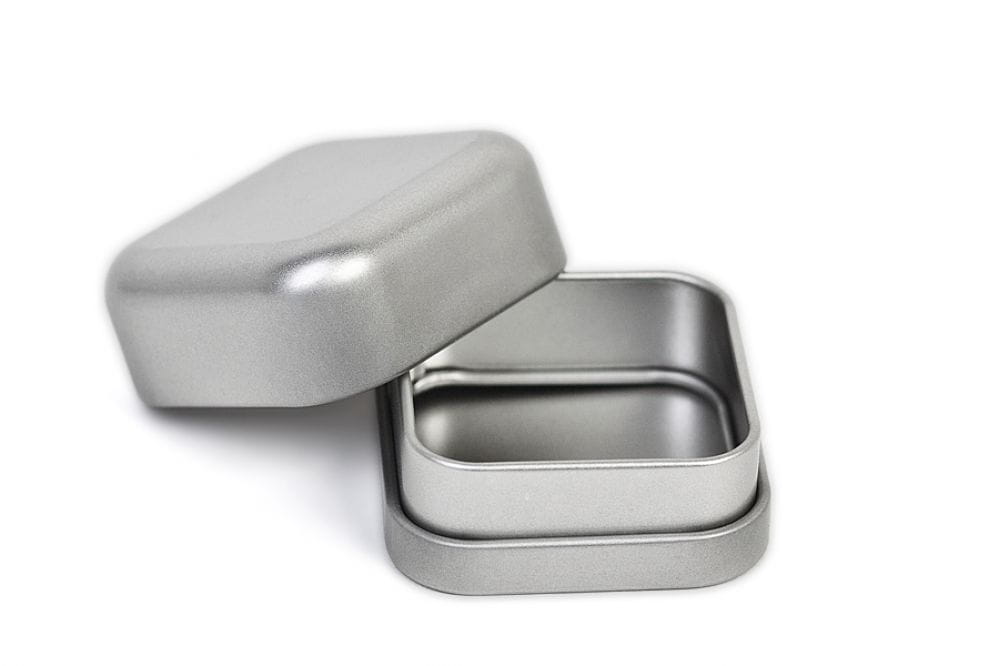 Geöffnete kleine quadratische Metalldose aus silberfarbenem Metall, mit leicht abgenommenem Deckel neben dem Korpus, auf weißem Hintergrund fotografiert.