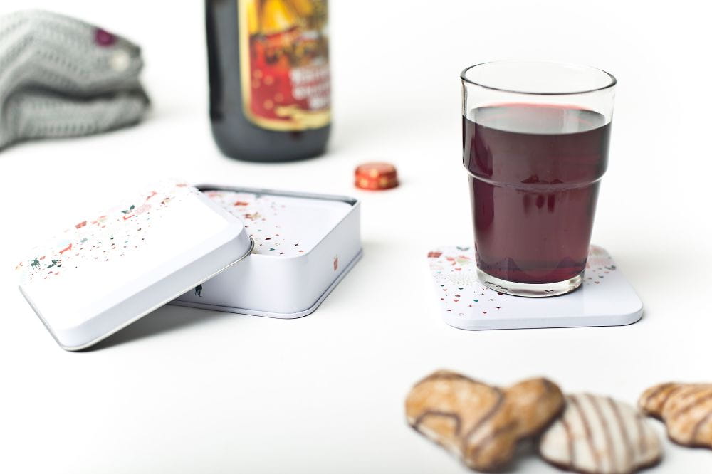 Ein Glas mit rotem Getränk auf einem weißen Dosenuntersetzer, daneben eine geöffnete Dose mit weihnachtlichem Design und ein paar Keksen. Der Hintergrund ist neutral.