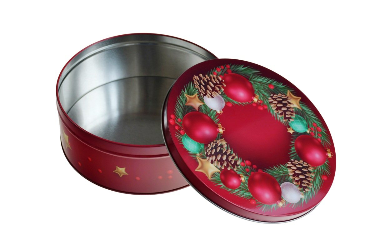 Geöffnete runde Blechdose mit weihnachtlichem Motiv, Deckel mit roten Kugeln, Sternen, Tannenzweigen und Zapfen danebenliegend, auf weißem Hintergrund fotografiert.