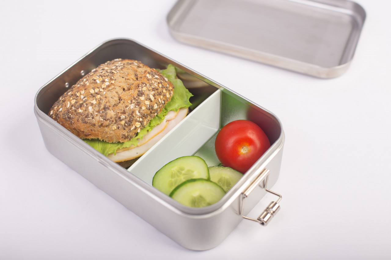 Edelstahl-Lunchbox Classic Maxi mit eingesetzter weißer Trennwand, gefüllt mit belegtem Brötchen, Tomate und Gurkenscheiben; Deckel im Hintergrund, neutraler Hintergrund.